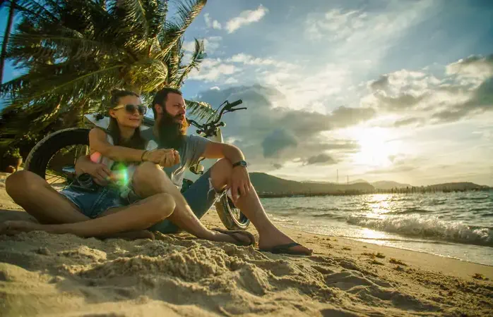 A couple - a bearded man and his handsome girlfriend - sitting on the beach leaned on motorbike, watching sunset at the sea